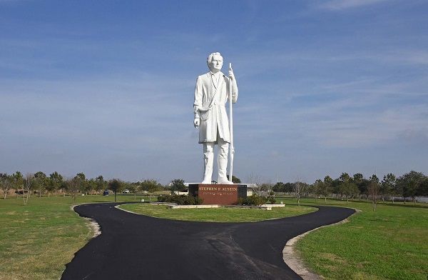 White statue on a brick pedestal in a grassy park under a blue sky, with a curving paved path leading to it
