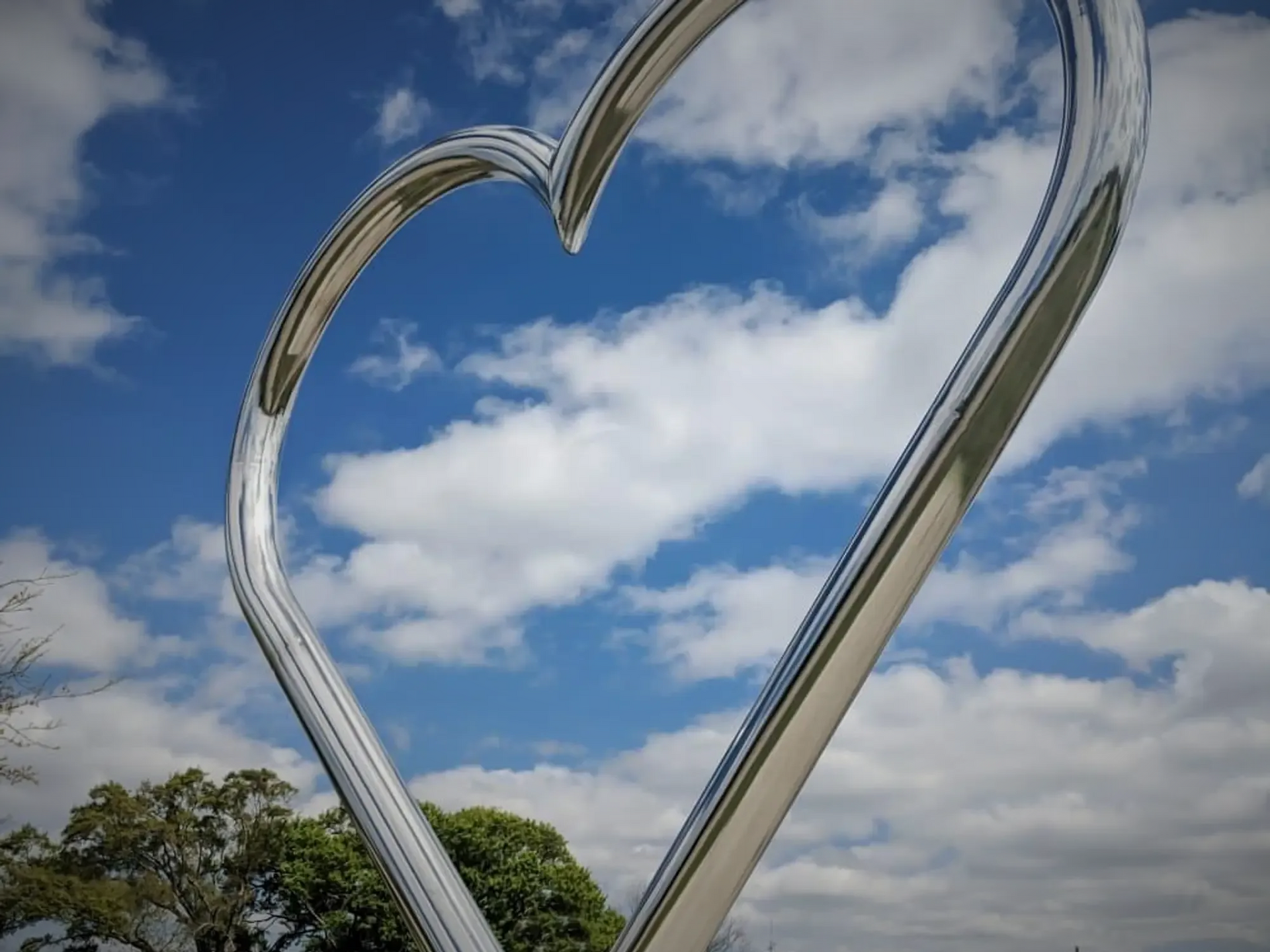 Large silver heart sculpture against a blue sky with clouds and trees below