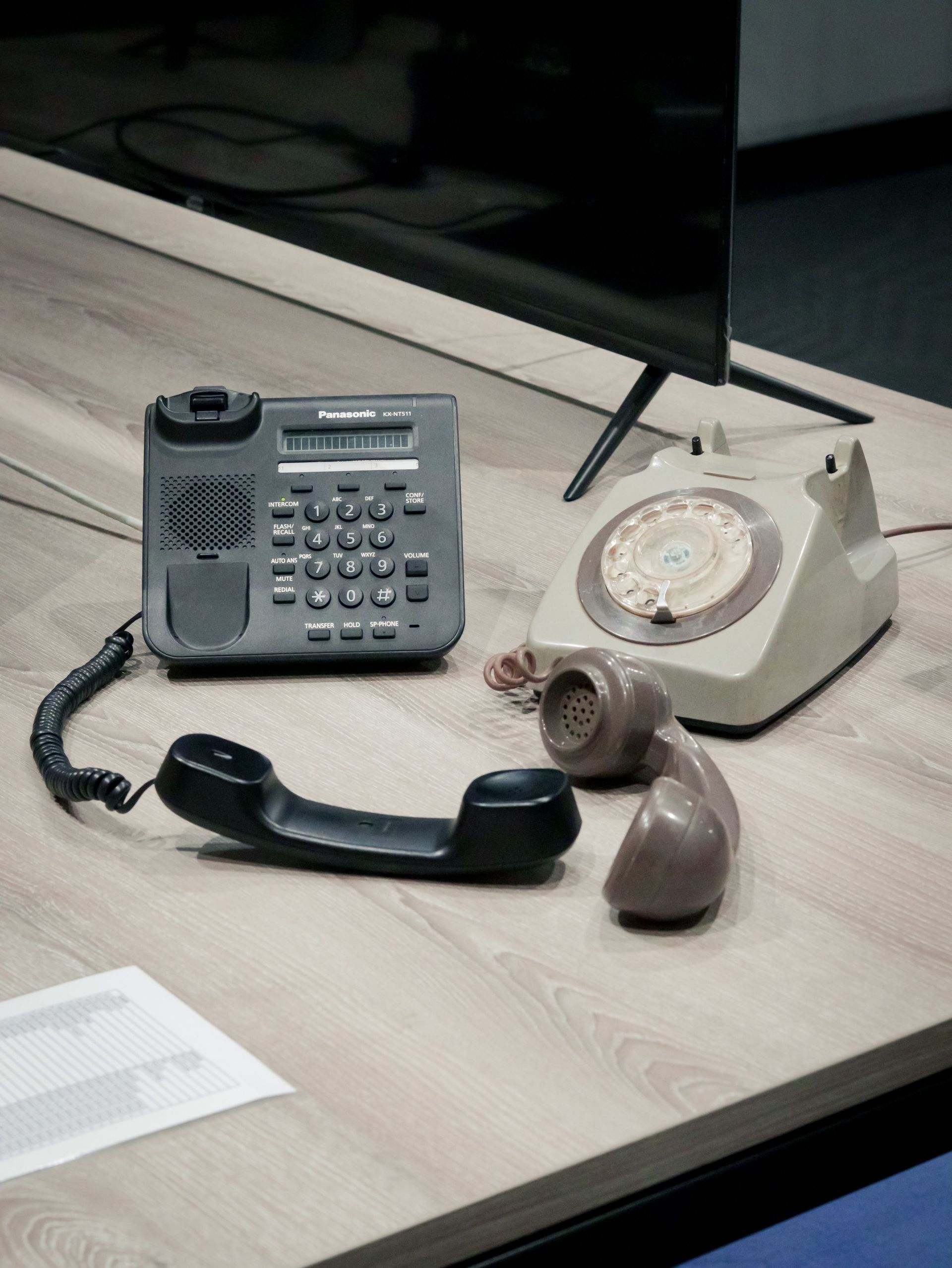 Desk with two office phones, one gray corded and one beige rotary, on a wooden tabletop.