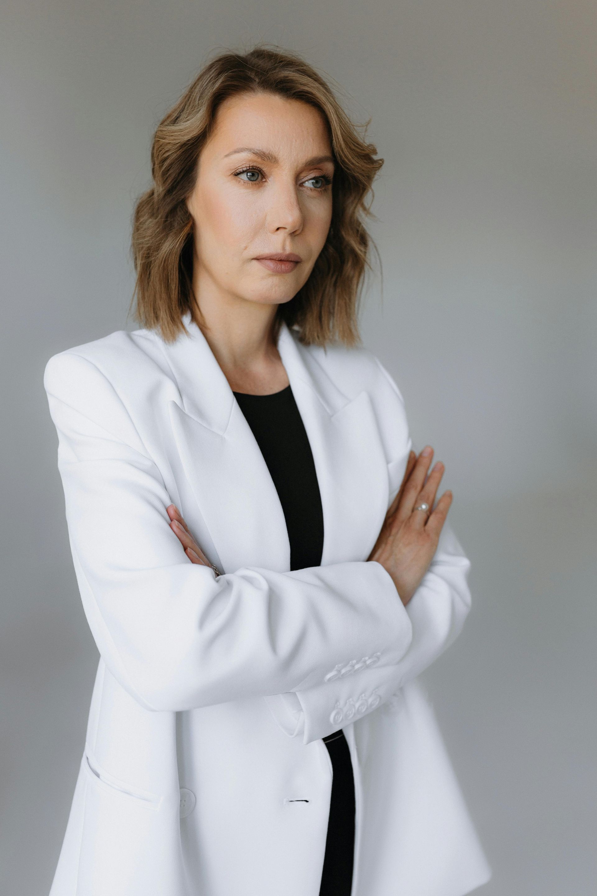 Woman in a white blazer with arms crossed, standing against a plain gray background.