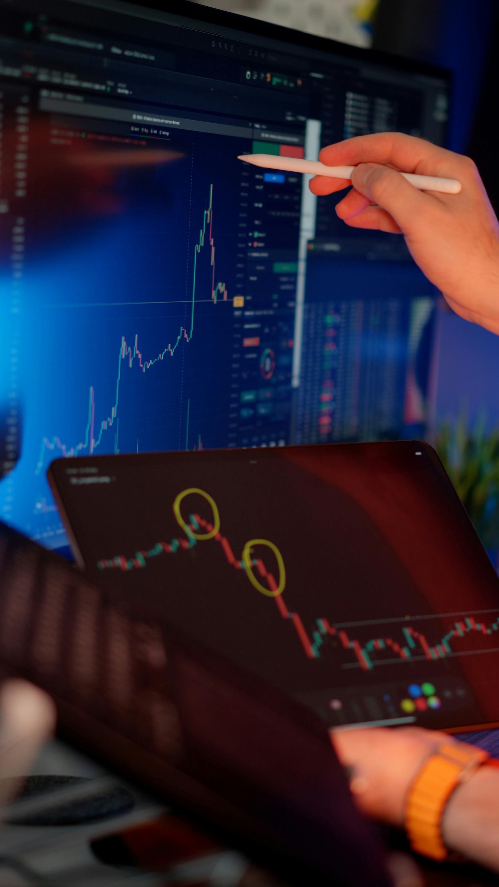 Hands analyzing stock charts on a tablet and monitor in a dimly lit trading setup.