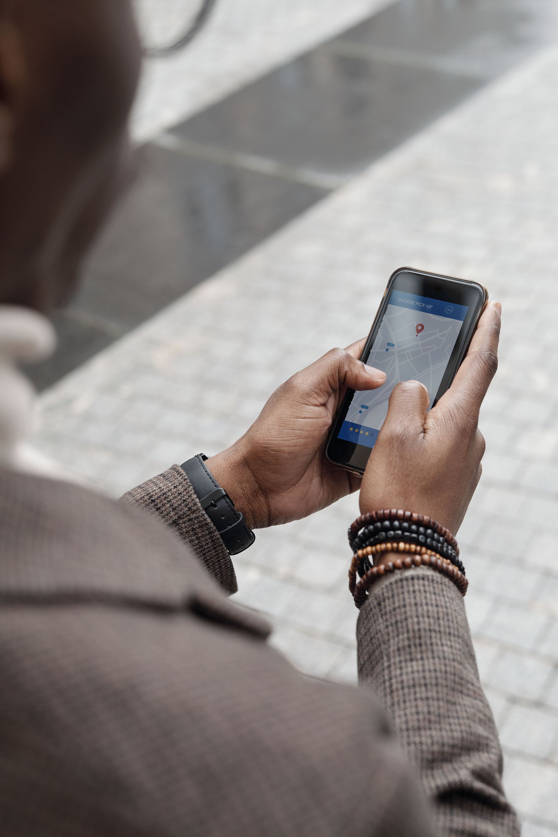 Person holding a smartphone and typing outdoors on a paved walkway, wearing a gray coat and beaded bracelets