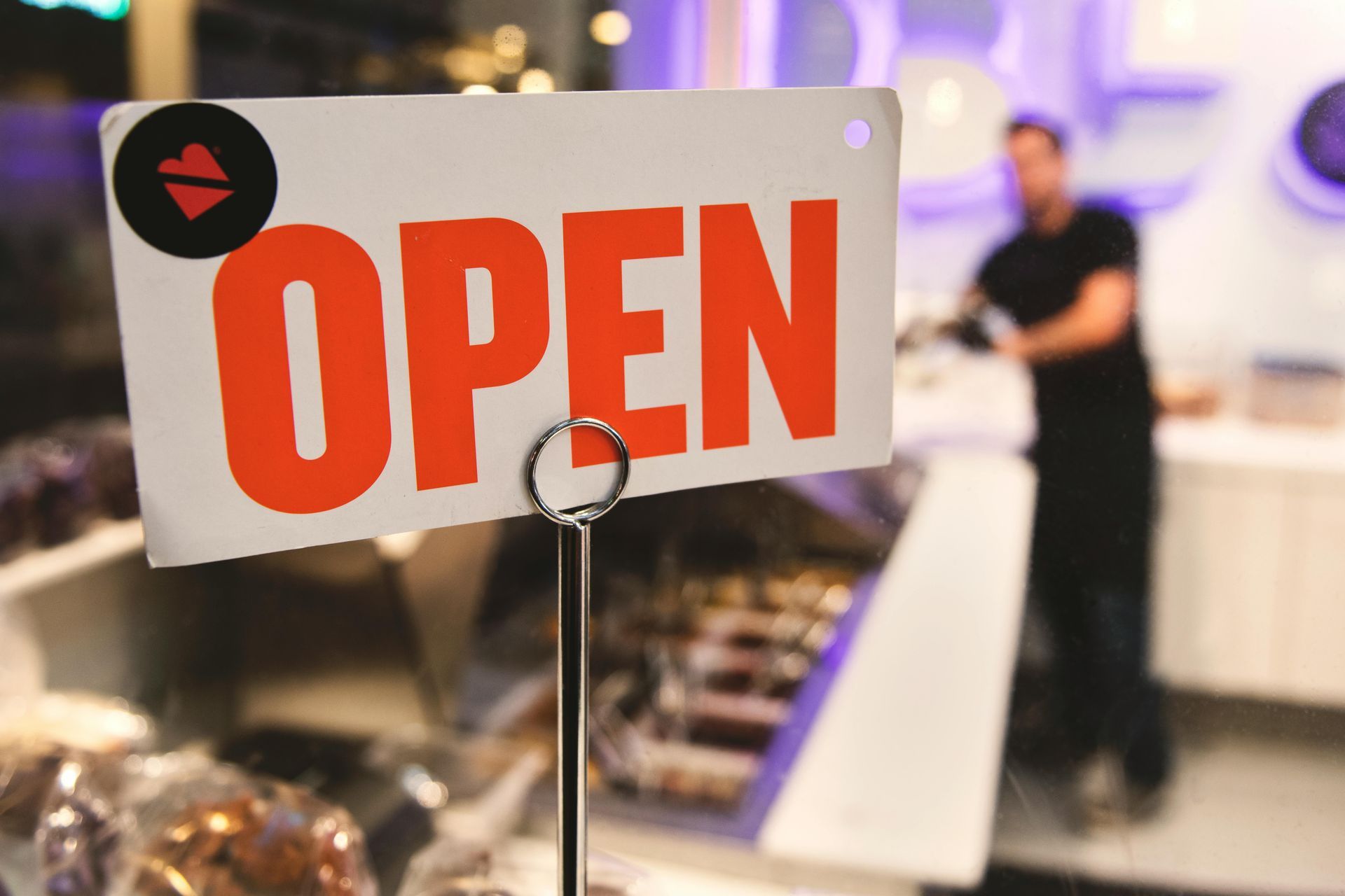 Open sign on a stand inside a shop, with a blurred person in the background.