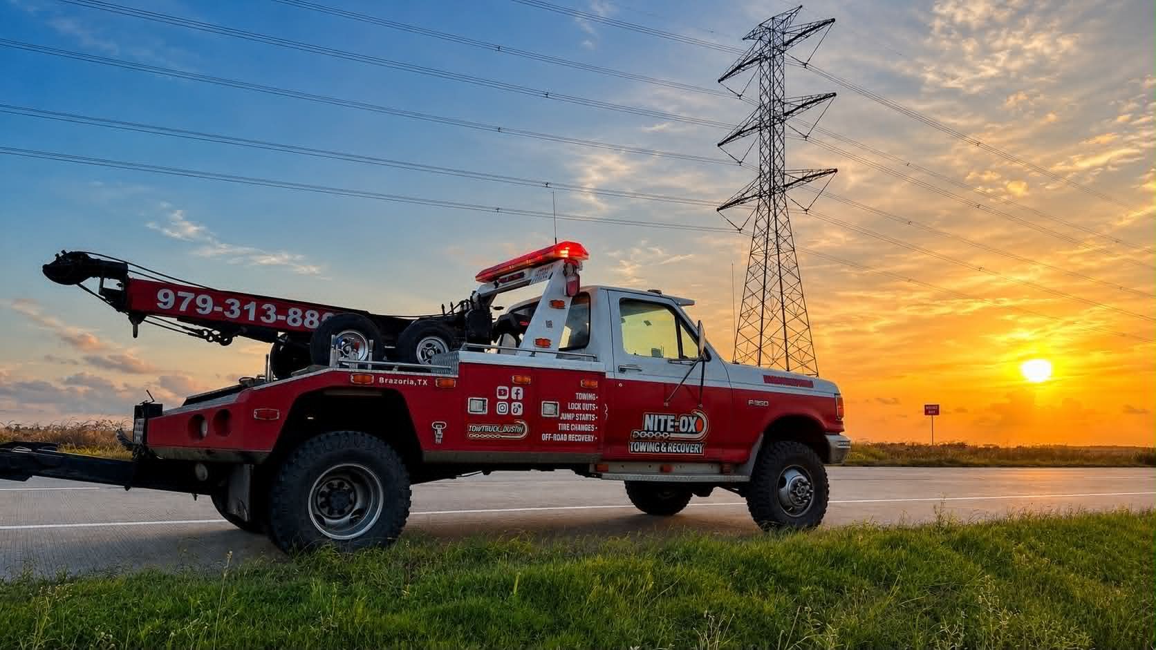 Red tow truck by a lake at sunset, with power lines silhouetted against the sky.