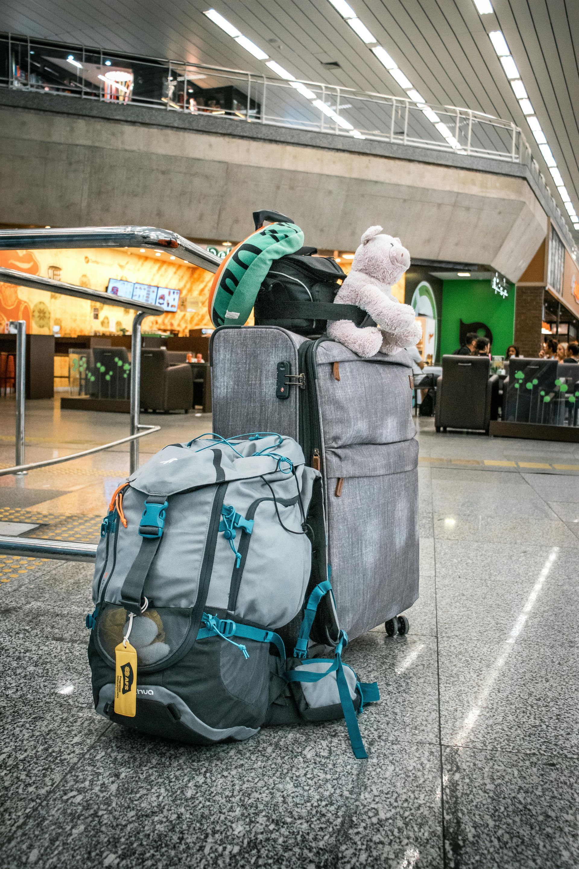 Mala e mochila empilhadas em um aeroporto. Um ursinho de pelúcia e um dinossauro de pelúcia verde estão em cima.
