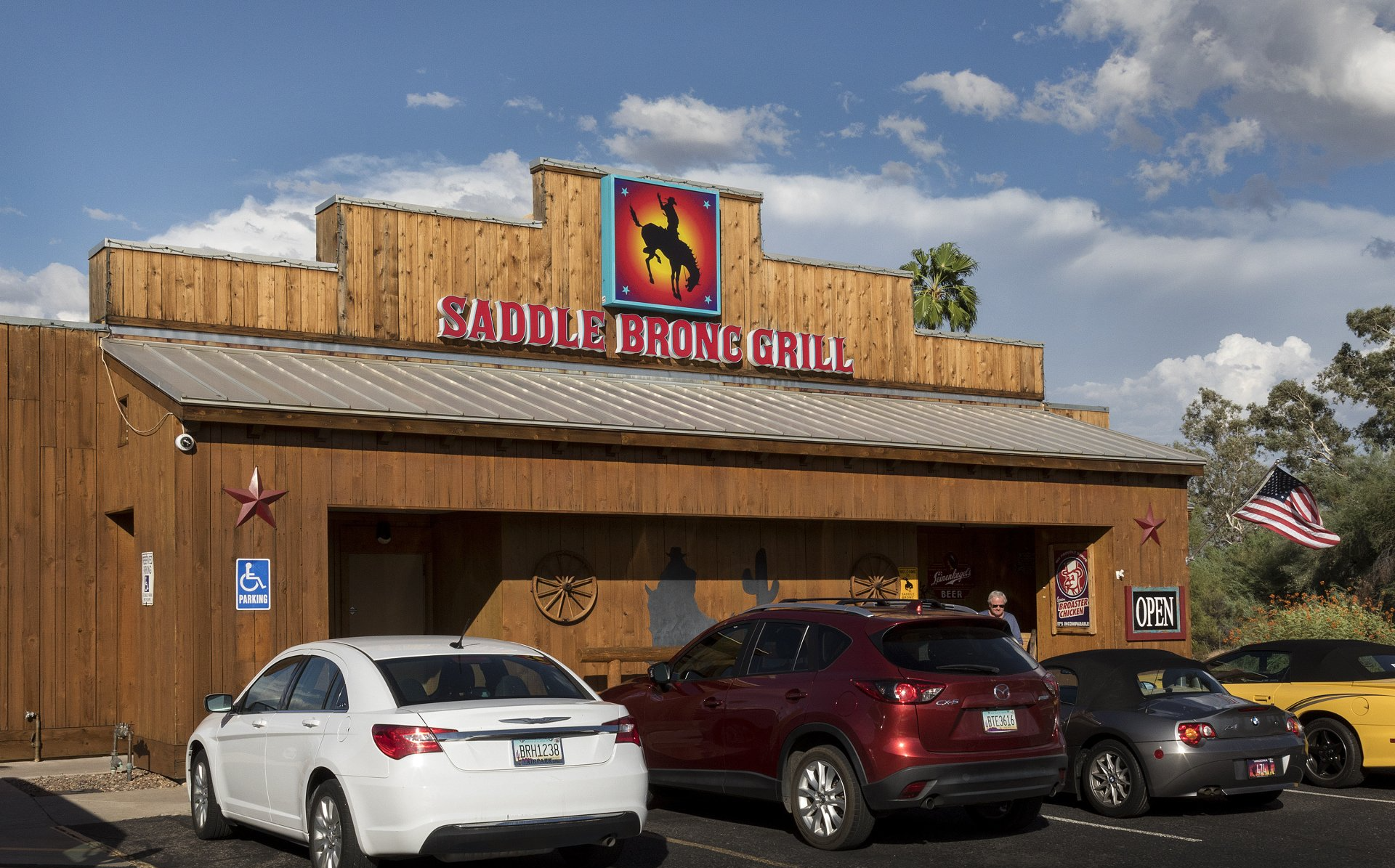 Saddle Bronc Grill, brown wooden exterior with cars parked in front, blue sky with clouds.