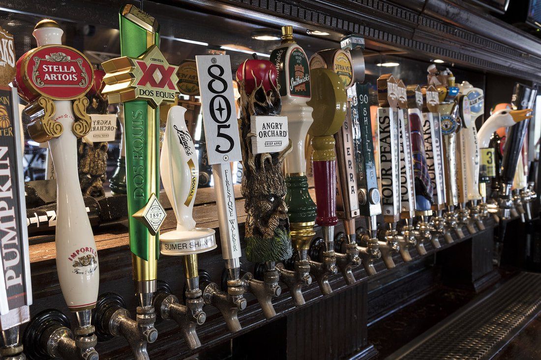 Row of beer taps at a bar, each with a unique handle. Wooden bar and background.