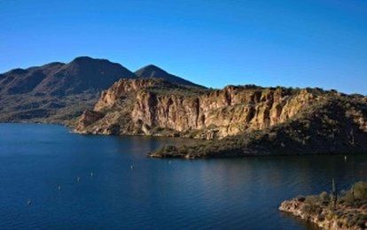 Lake with mountains and cliffs on a sunny day, clear blue sky, and desert vegetation.