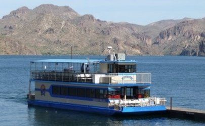 A two-story blue and white cruise boat docked at a pier on a lake with mountains in the background.