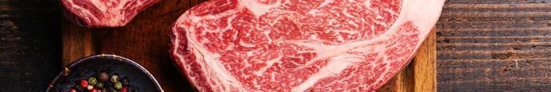 Two thick cuts of raw steak with marbling, on a wooden cutting board, alongside a small bowl of peppercorns.