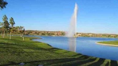 Lake with fountain, grassy shore, trees, and buildings under a blue sky.