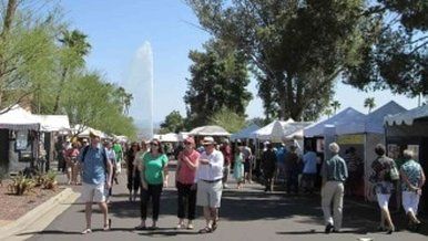 Crowd at outdoor art fair; people walking, browsing white vendor tents, tall fountain in distance.
