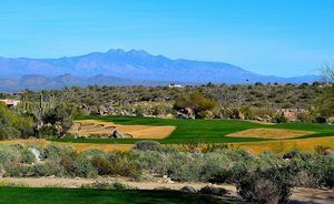 Golf course with green fairways, desert shrubs, and mountains in the distance under a blue sky.