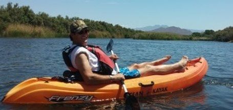 Man relaxing in an orange kayak on a lake, wearing a hat and life vest.