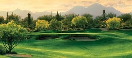 Green golf course with trees, sand traps, and mountains in the background under a golden sky.