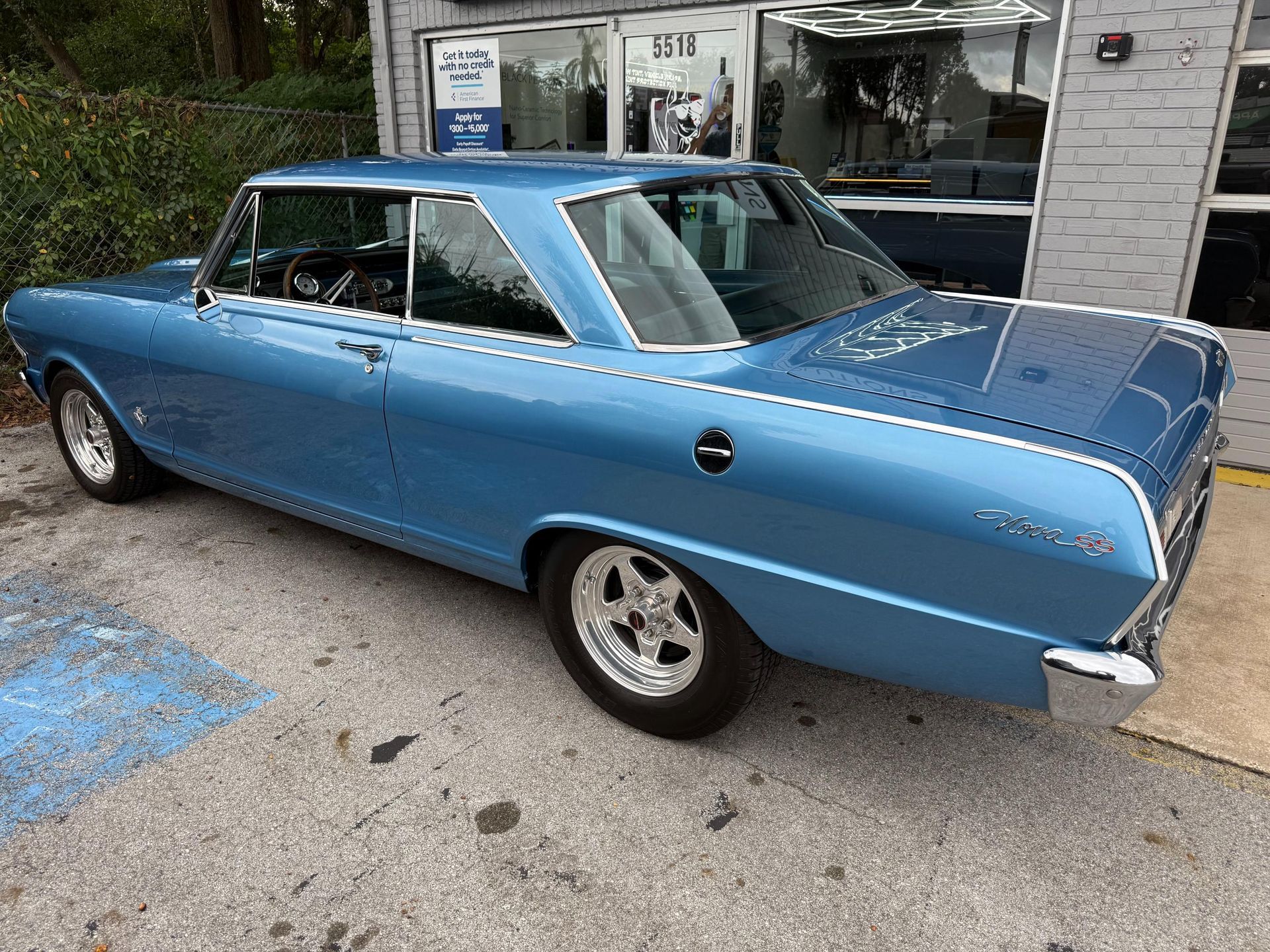 Blue 1965 Chevrolet Nova coupe parked outside a shop. Chrome trim, mag wheels.