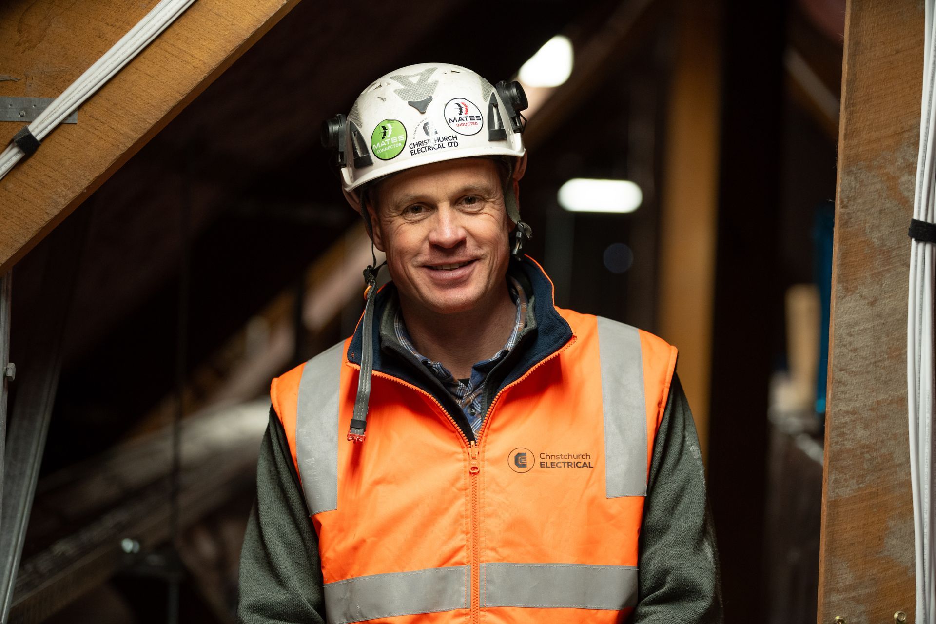Electrician in hard hat and hi-viz vest  in roof space at CBHS smiling.