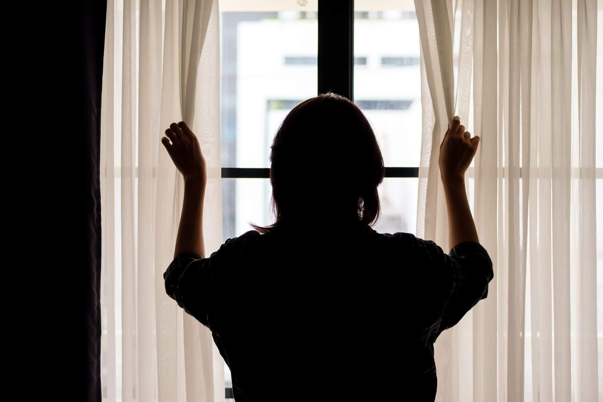 Silhouette of a person opening curtains, looking out a window at daylight.