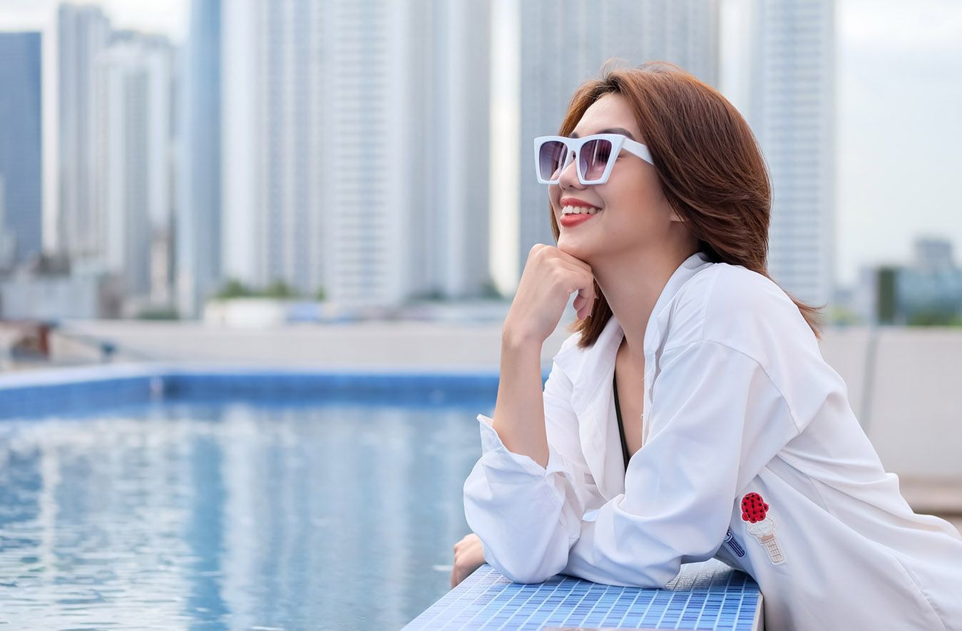 Woman in white sunglasses and shirt smiling by a rooftop pool with city skyscrapers in the background