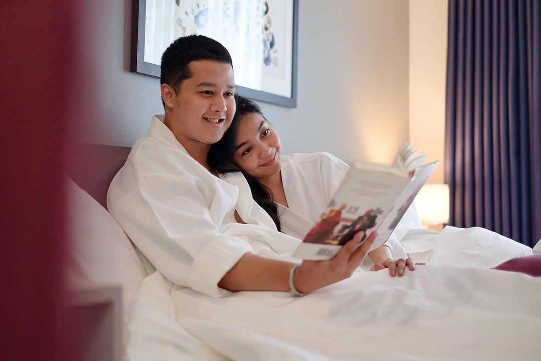Two people in white robes sit on a bed in a bedroom, smiling while reading a book together.