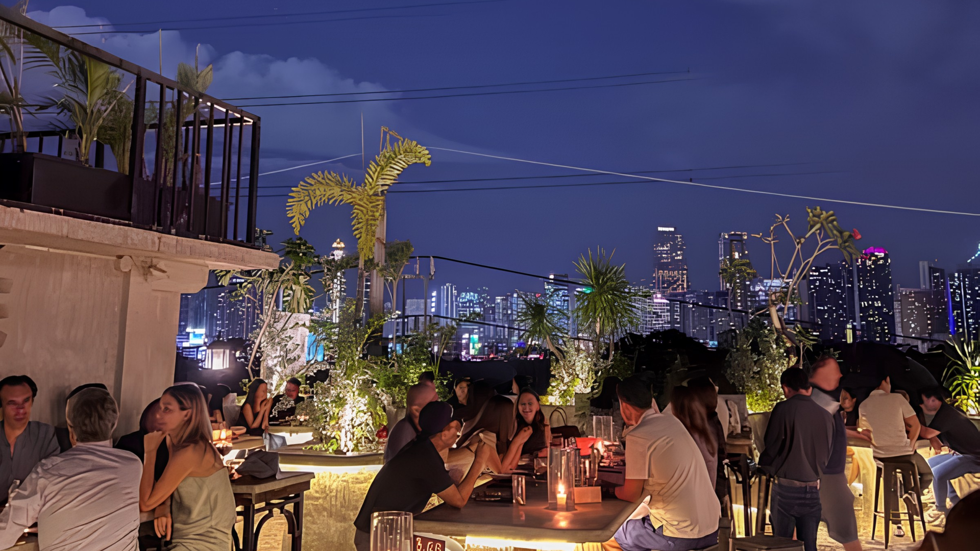Rooftop dinner party at dusk with people seated at tables, city skyline and purple sky in the background.