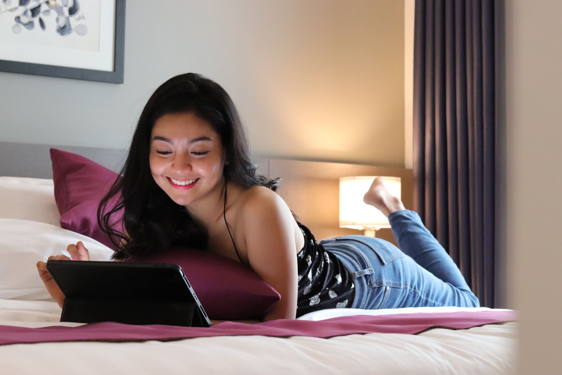 Smiling traveler lying on a bed with plum accents while using a tablet in a guest room at The Belamy House
