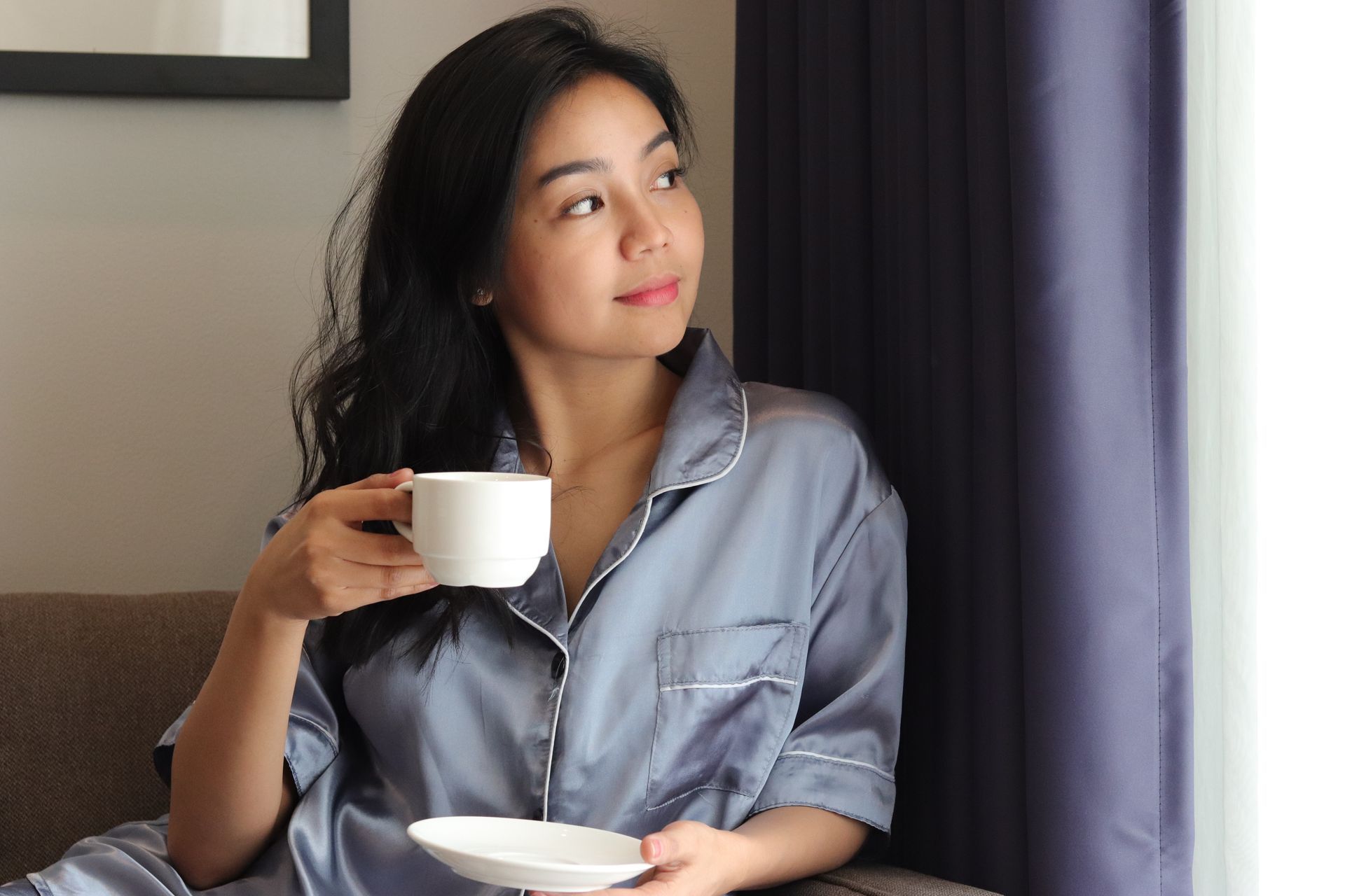 Relaxed guest in satin pajamas sipping morning coffee by the window of a room at The Belamy House in Makati