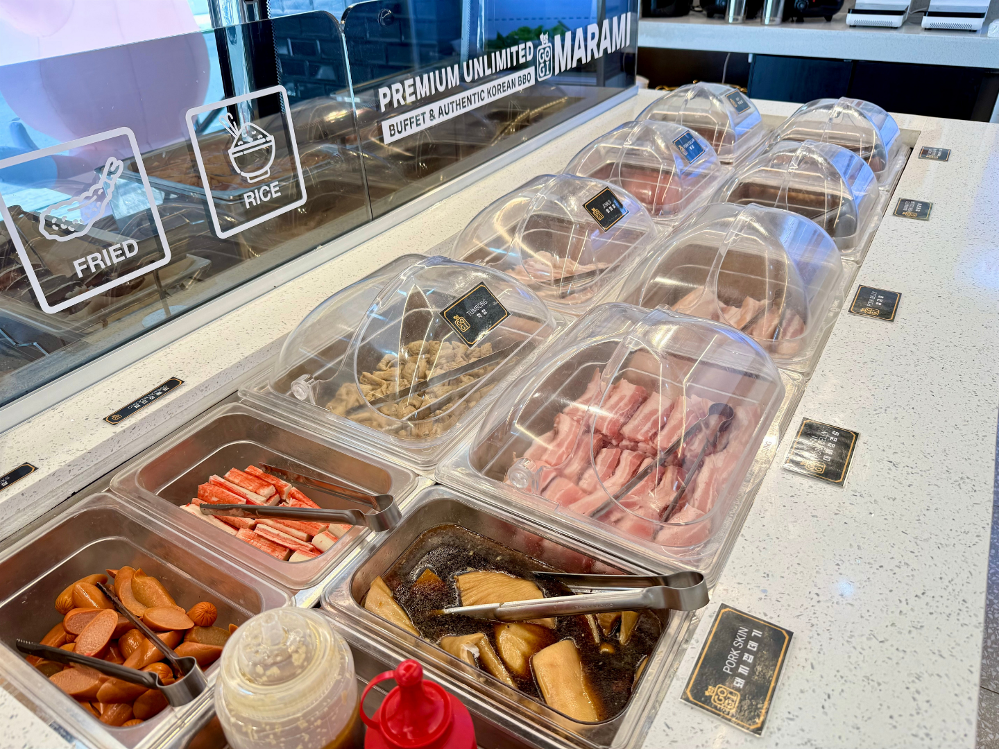Self-serve ice cream topping bar with covered bins of candy, fruit, and sauces under a glass display