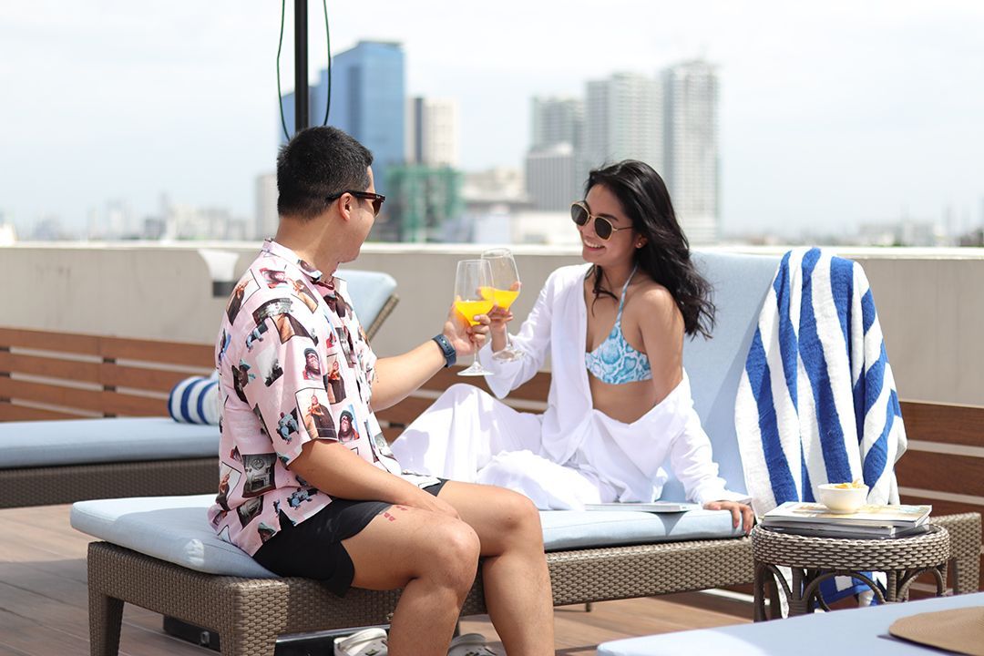 A couple in sunglasses cheers with glasses of orange juice while reclining on poolside lounge chairs on a city rooftop.