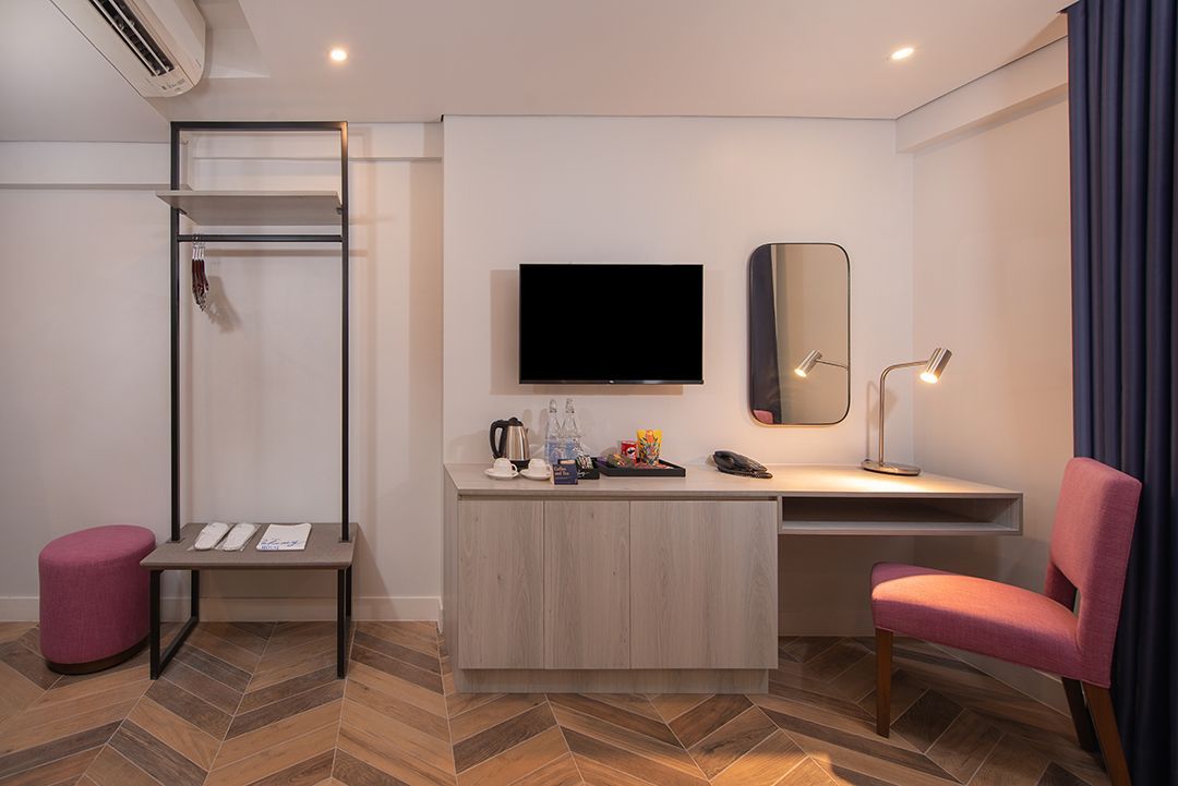 Modern hotel room with bed, desk, wall TV, mirror, pink chair, and open closet in soft neutral tones