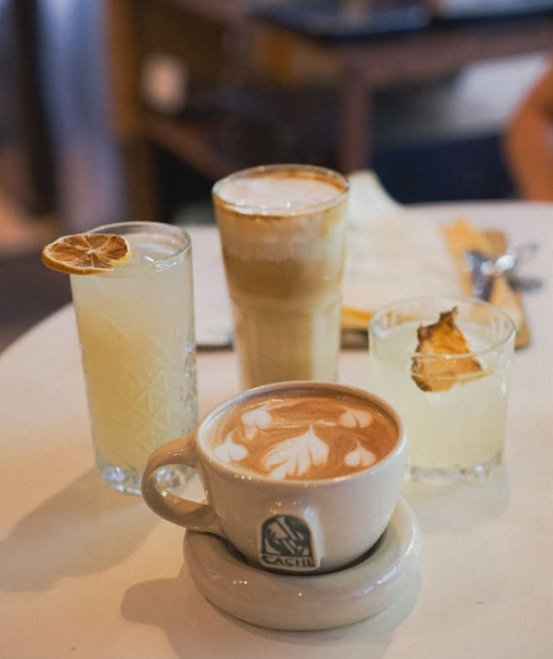 Four beverages: latte, iced drink, two cocktails on a white table. Warm lighting, coffee cup has latte art.