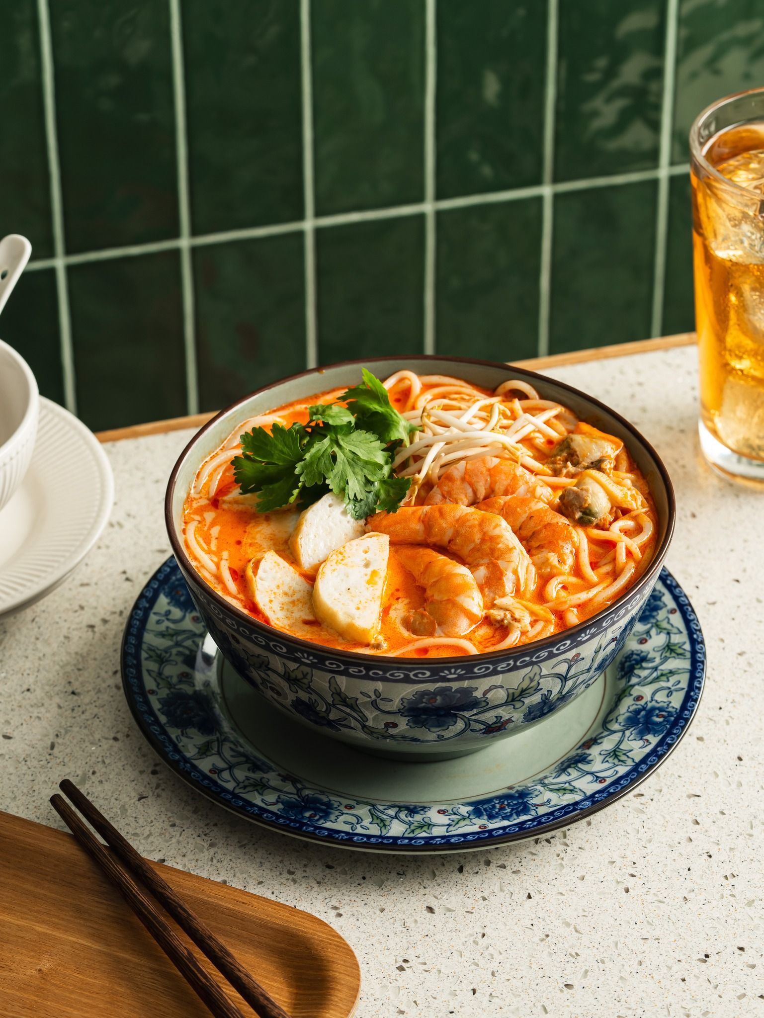 A bowl of laksa noodles with prawns and herbs, served on a patterned plate next to chopsticks and a glass of tea.