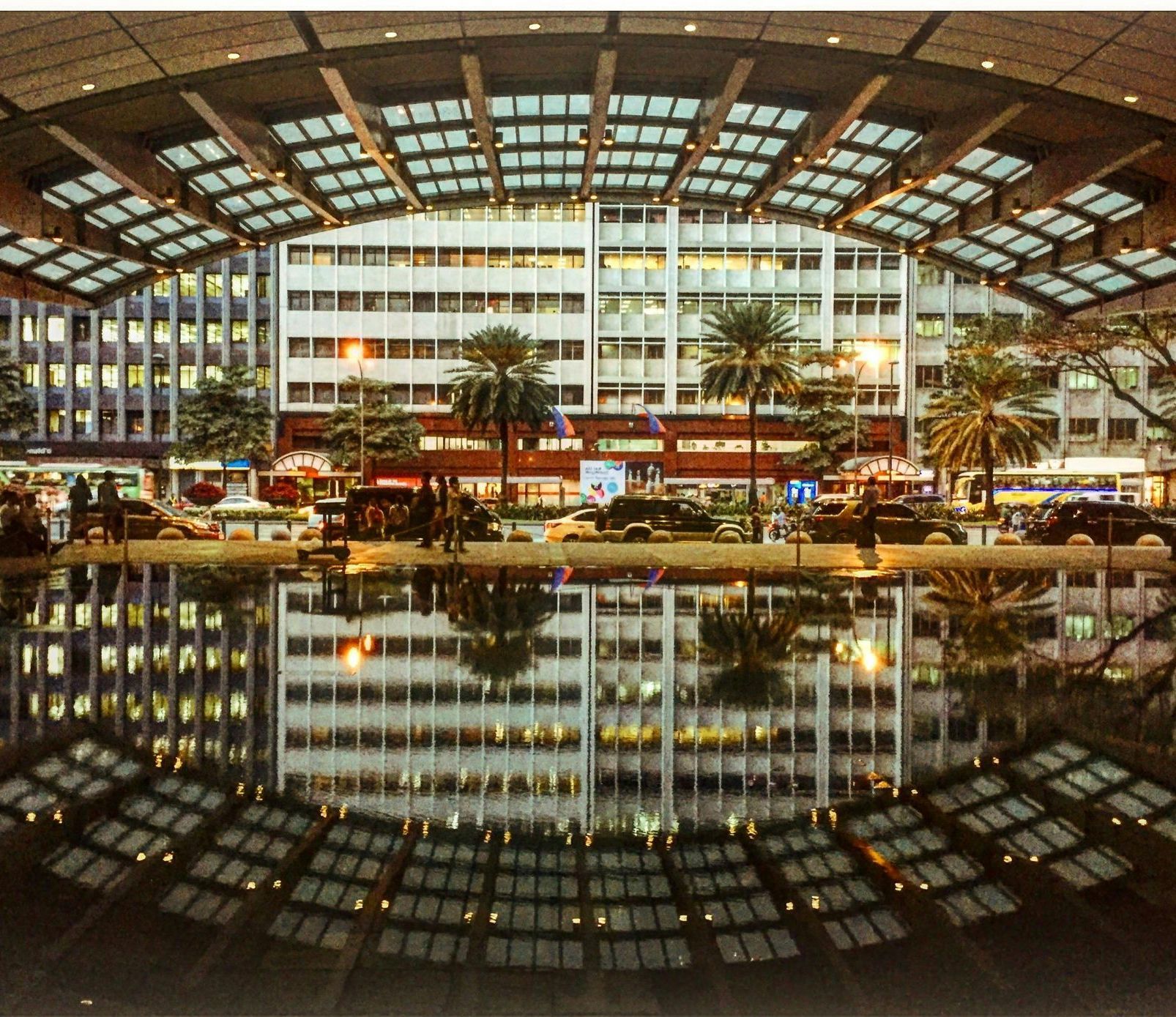 Buildings and arching roof reflected in water; street with cars, palm trees, and lights at night.