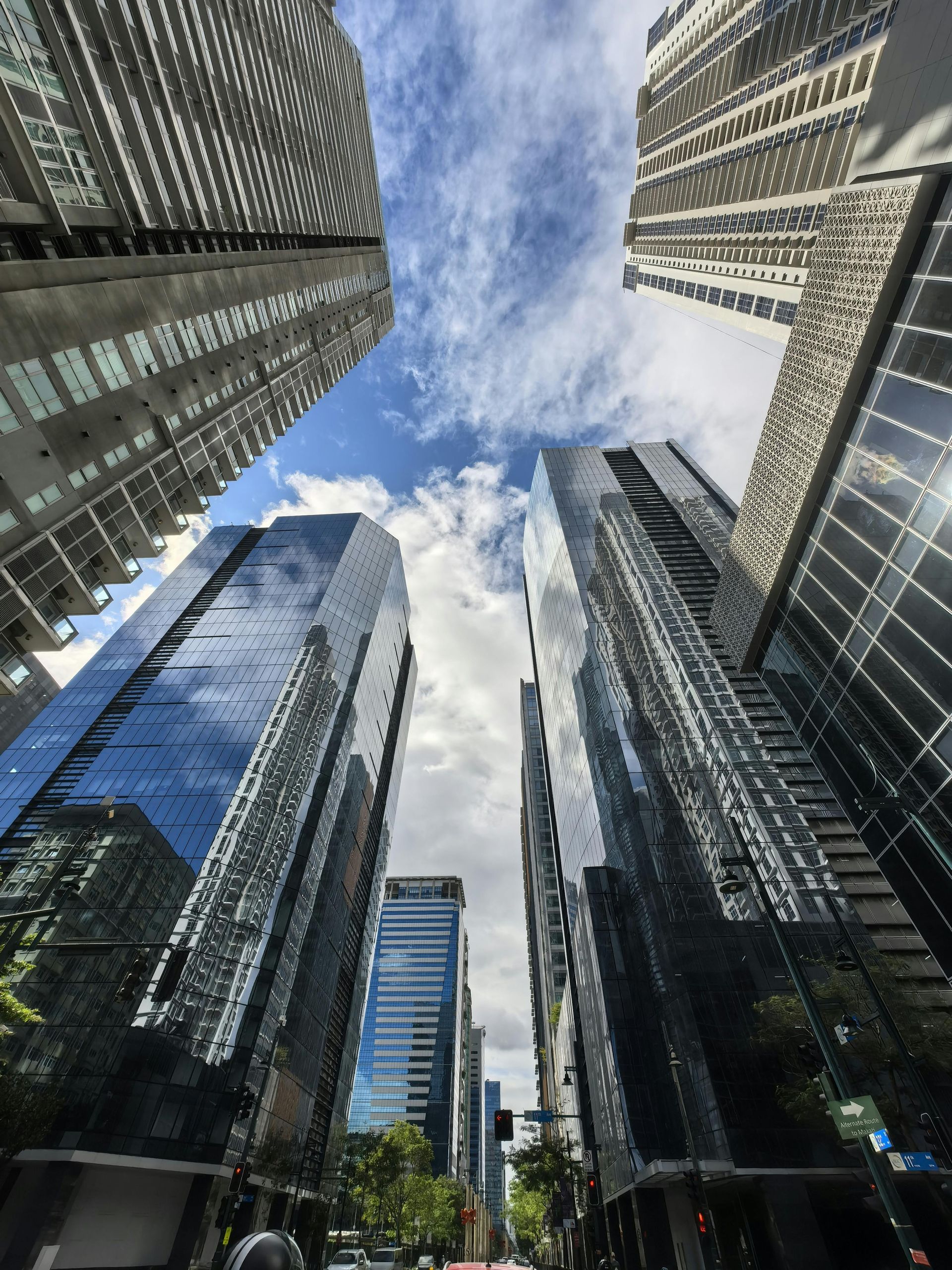 Looking up at modern skyscrapers in a city, blue sky peeking through, reflective glass exteriors.