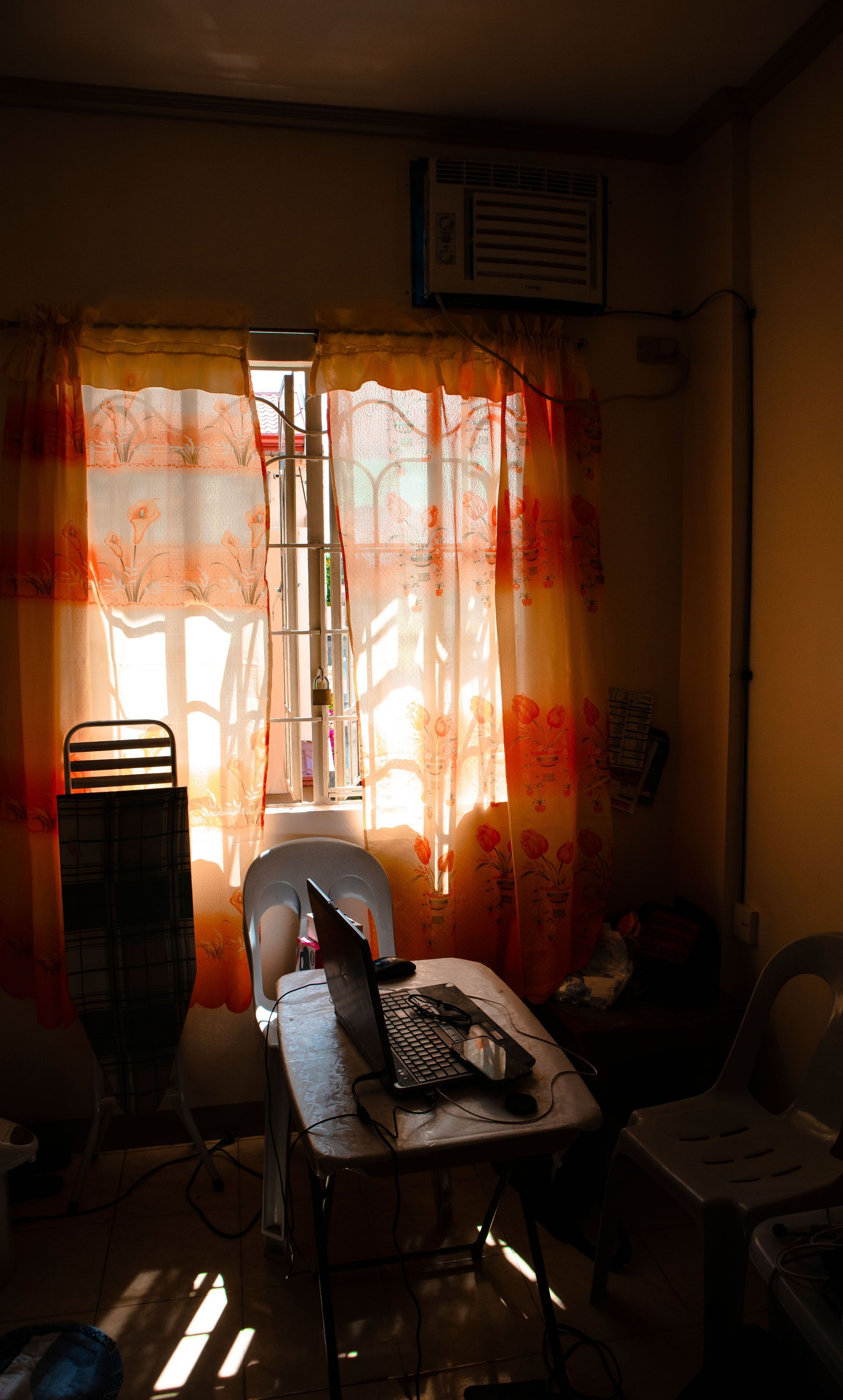 Dim room with sunlight through orange curtains, an ironing board, and a chair by the window.