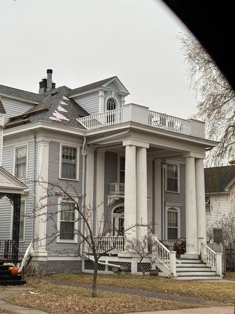 A light blue two-story historic house with a prominent white columned porch and a roof terrace, set against a gray sky.