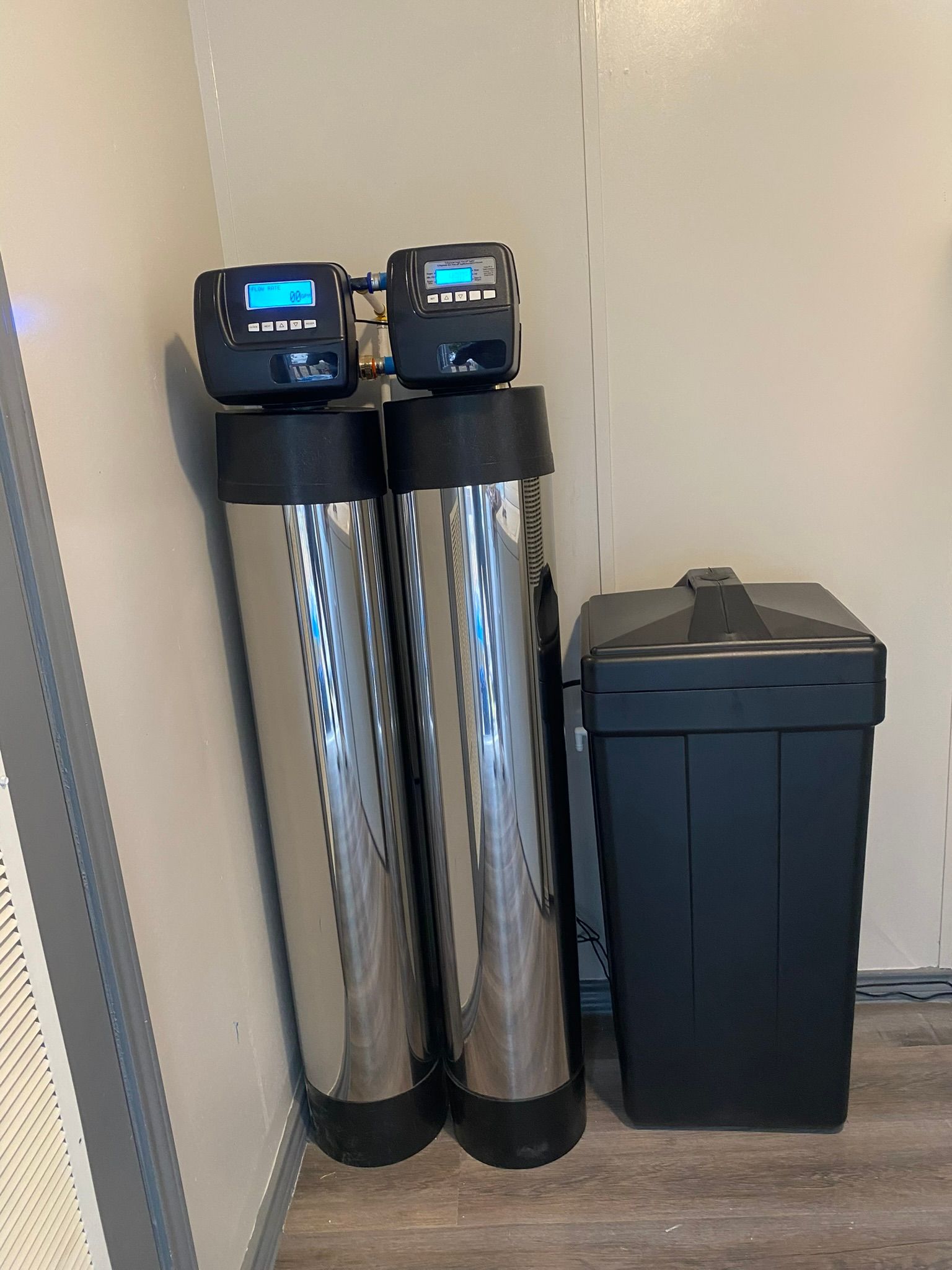 Two tall, metallic water filtration tanks with digital displays and a black salt tank in a utility room.