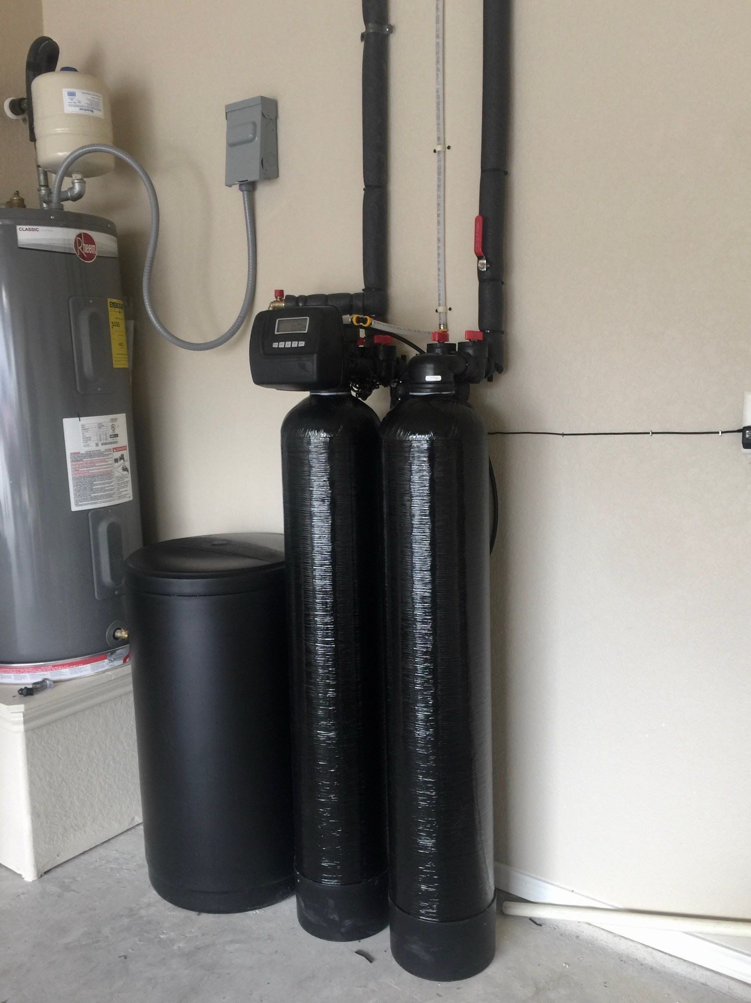 Water softener system in a garage with a water heater. Black tanks, pipes, and a pressure tank.