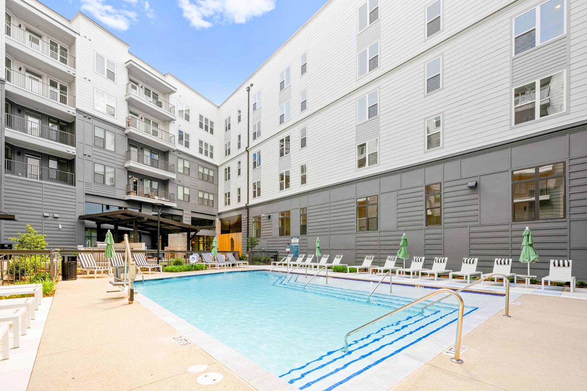 Swimming pool surrounded by a multi-story apartment building, white lounge chairs, and green umbrellas.