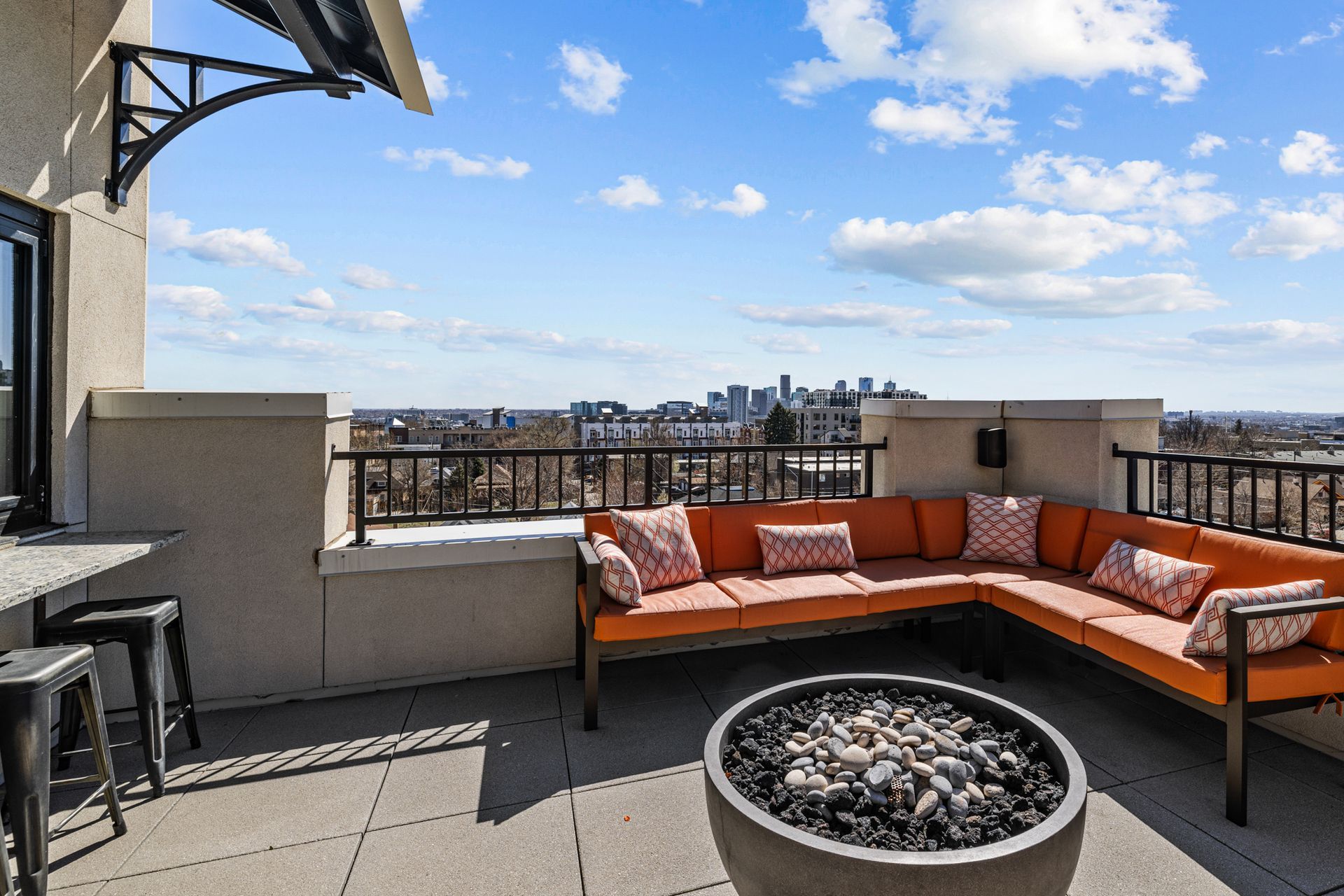 Rooftop patio with orange sectional sofa, fire pit, and cityscape view under a blue sky.