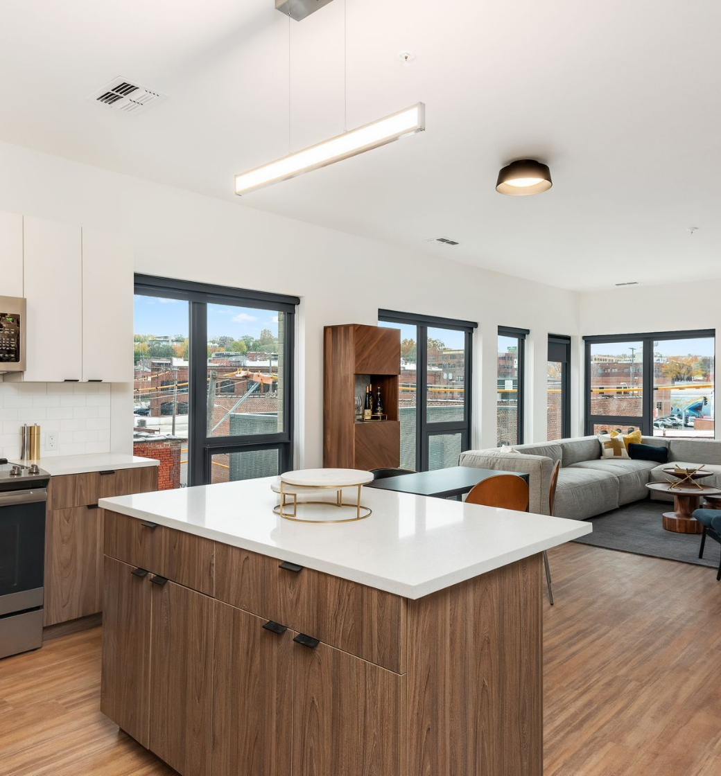 Modern open-plan kitchen and living room with wood floors, large windows, white island, and gray sectional sofa