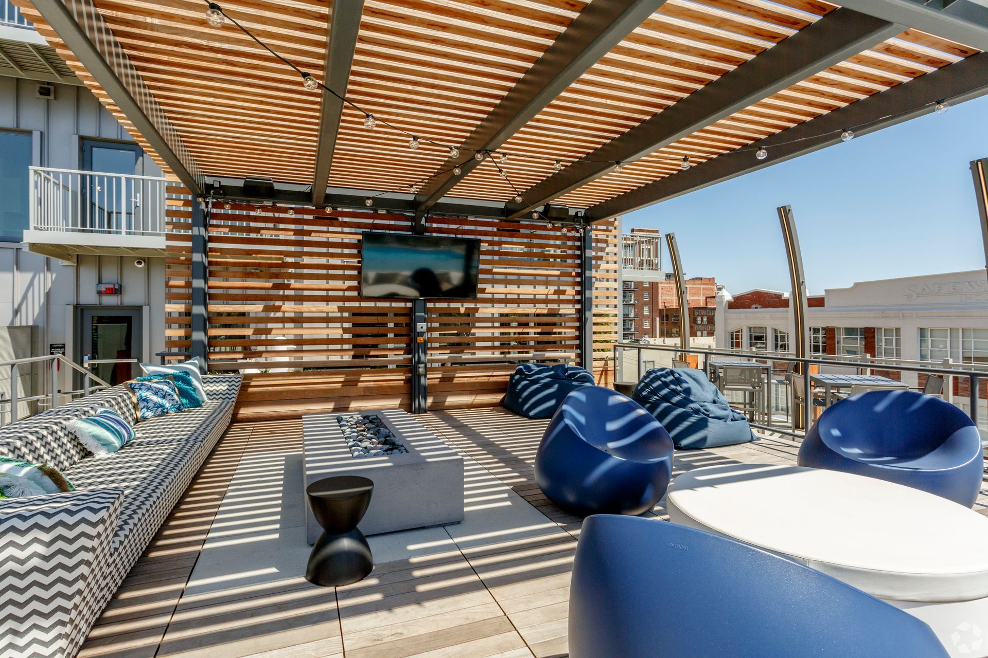 Rooftop lounge with slatted wooden shade, seating, fire pit, TV. Blue accents, bright sunlight.