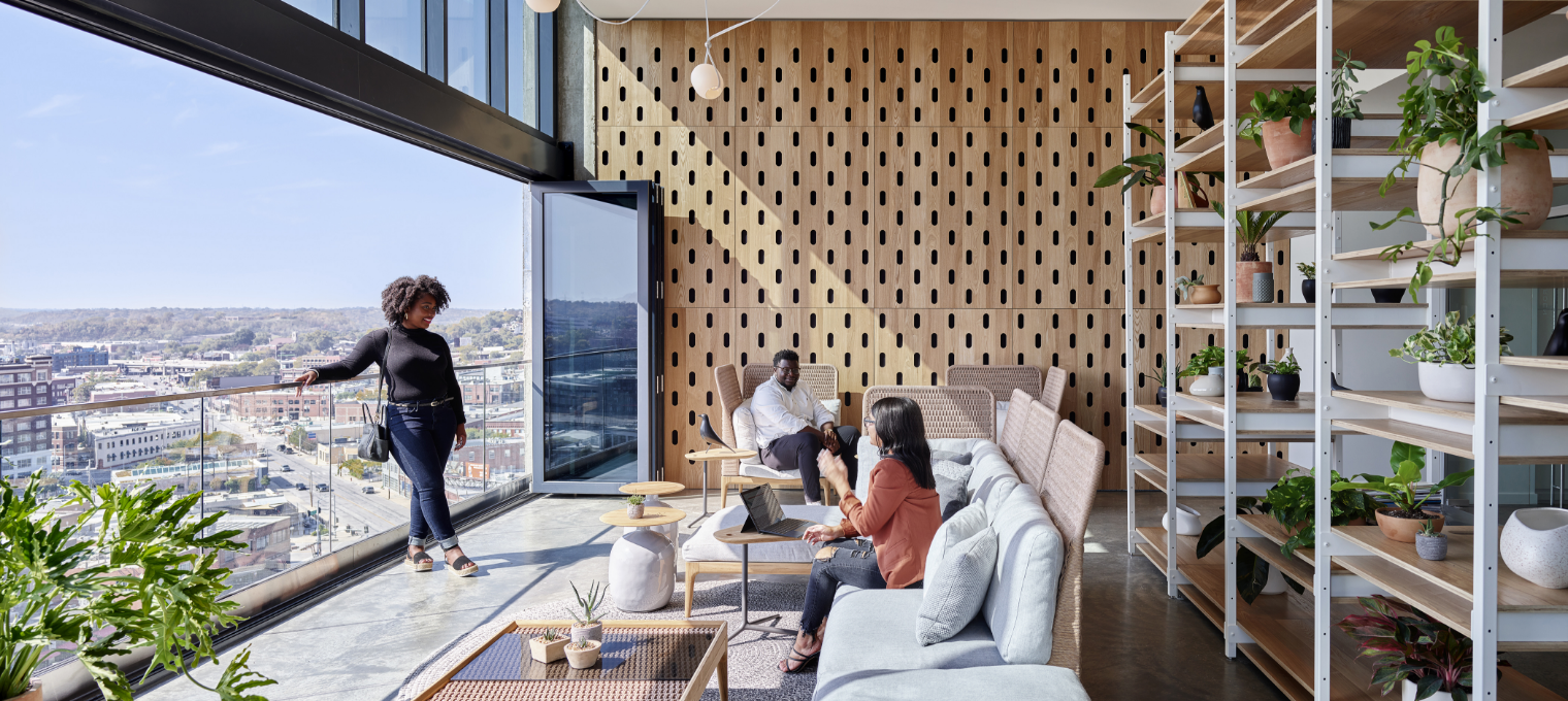 A modern rooftop lounge with city view. People on sofa, woman on balcony, wood paneling, plants. Sunny day.