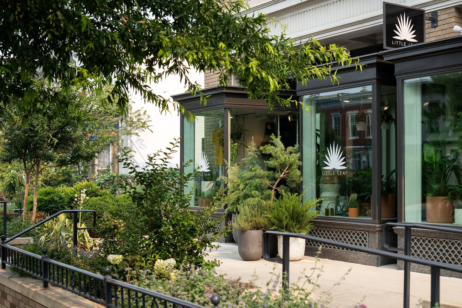 Exterior of a plant shop with large windows and various potted plants. Black awning and a black metal railing.