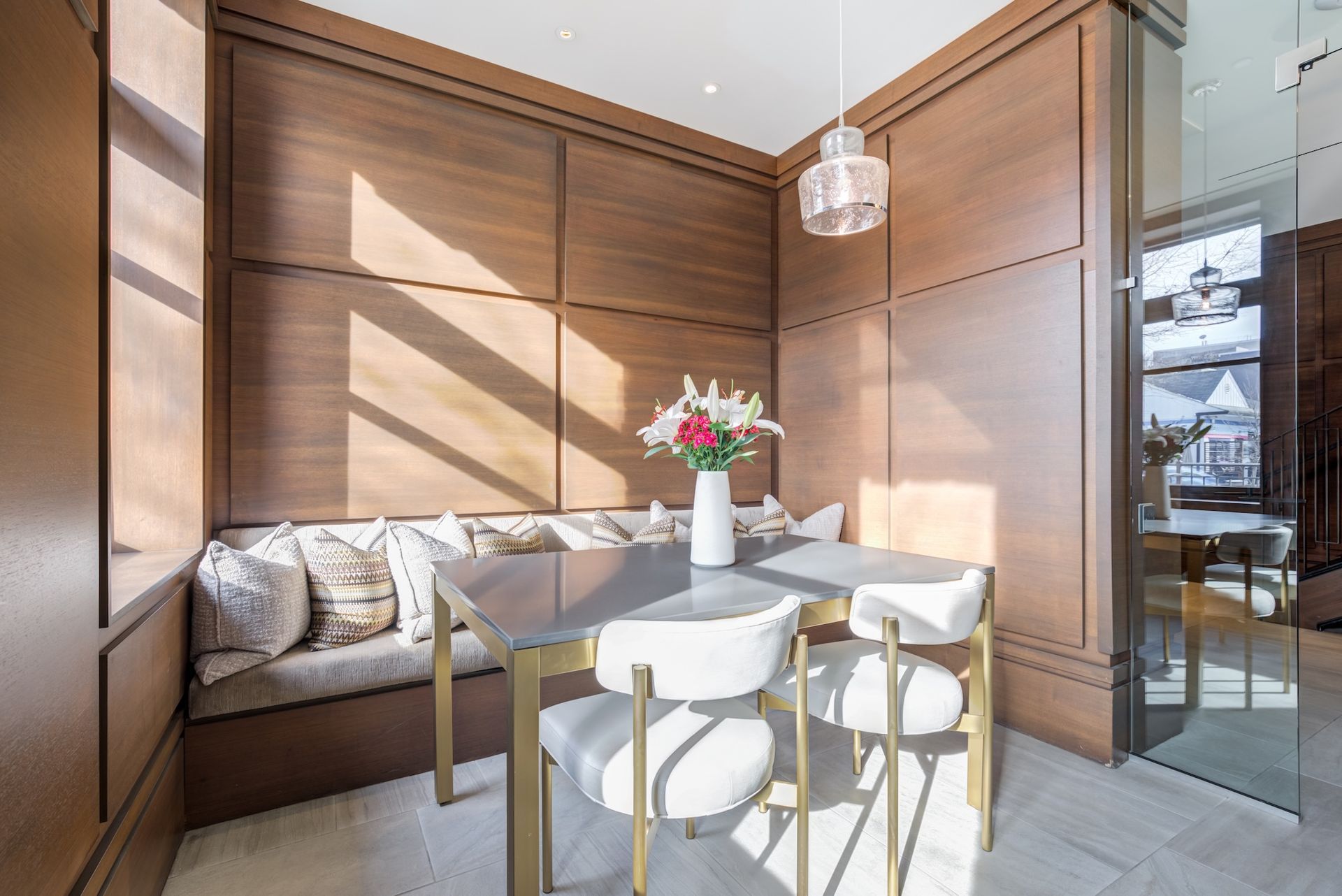 Modern sunlit dining nook with wood-paneled walls, a bench, small table, chairs, and a vase of flowers