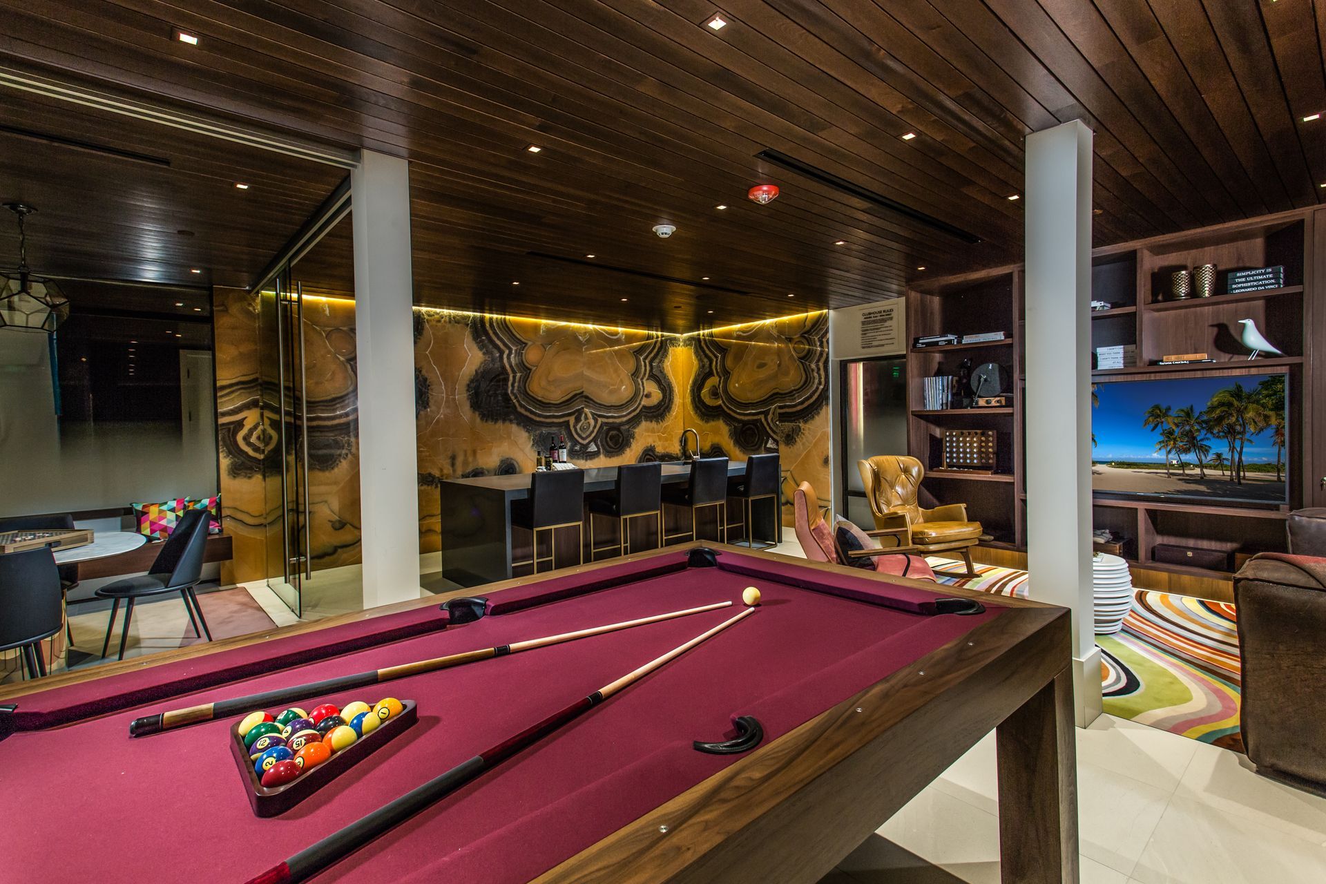Pool table in a luxurious room with a bar, bookshelves, and wood-paneled ceiling.