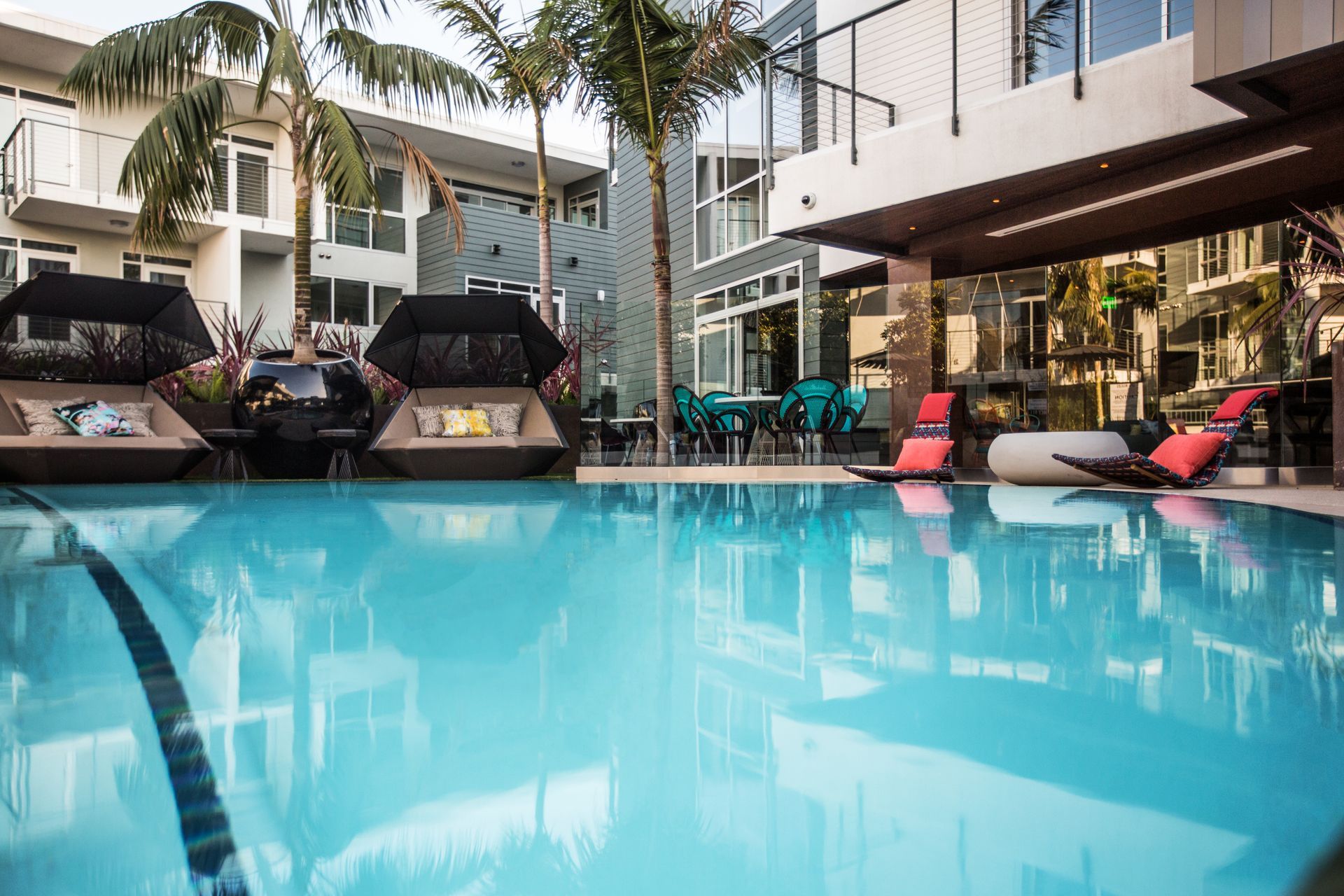 Swimming pool with lounge chairs and buildings. Palm trees and blue sky are visible.