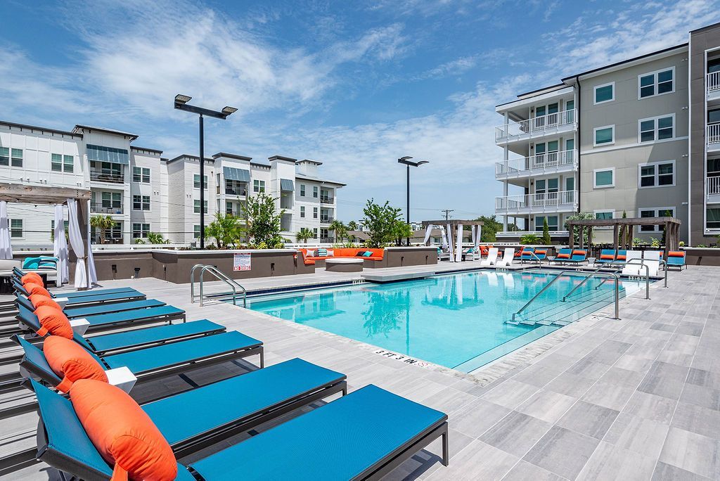 Swimming pool with lounge chairs, surrounded by apartment buildings under a blue sky.