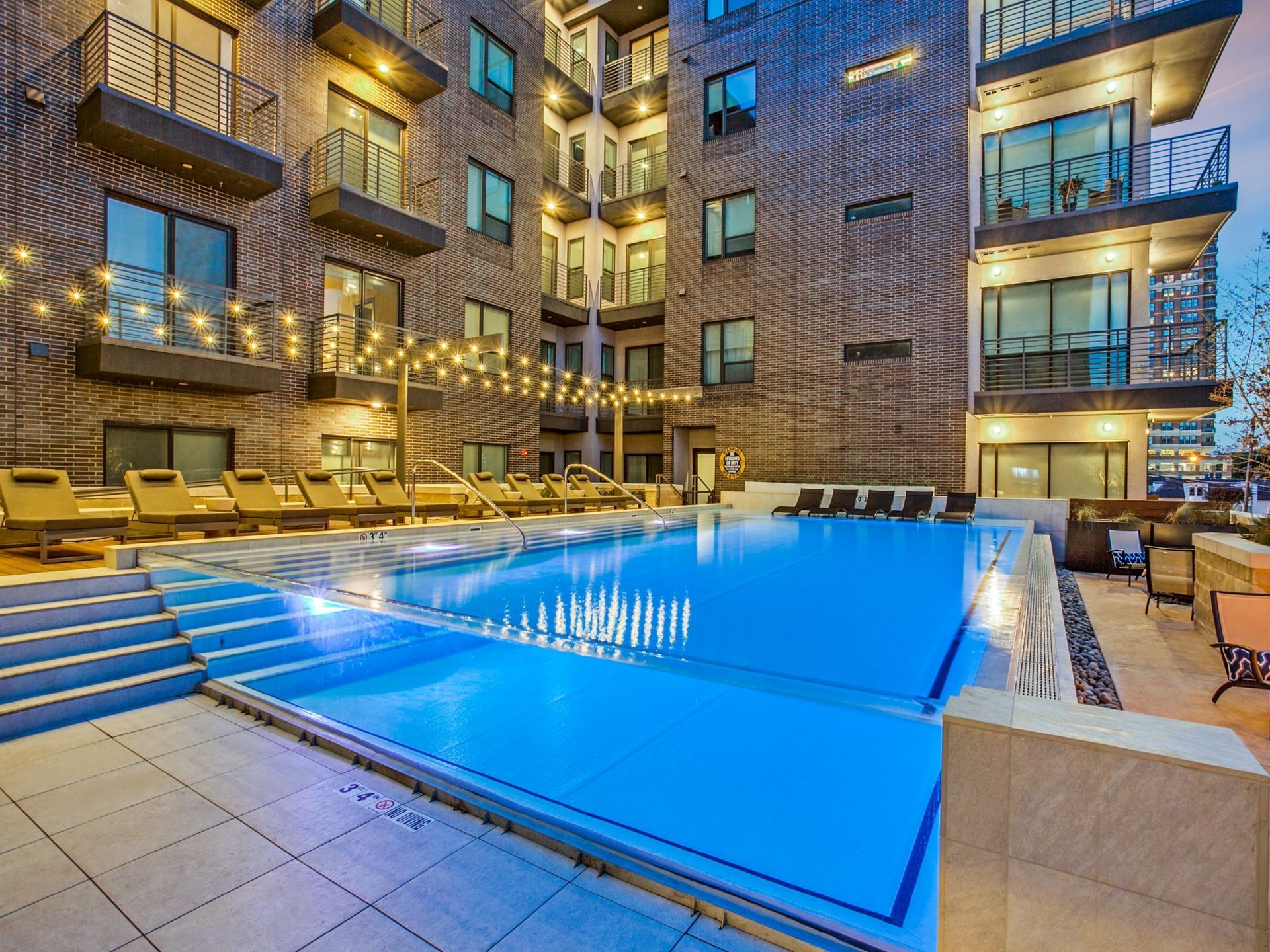 Outdoor swimming pool with lounge chairs, surrounded by a modern building with balconies and string lights.