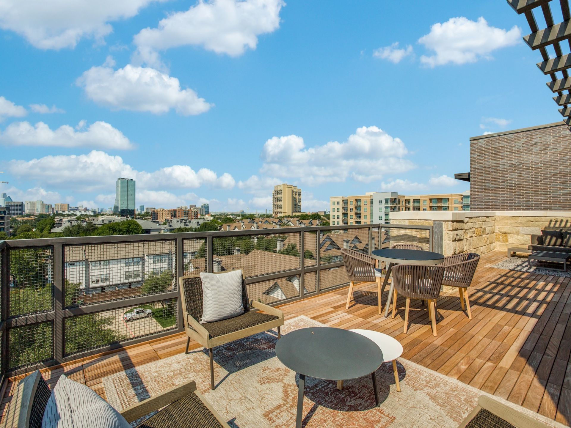 Rooftop patio with seating, cityscape view under a blue sky with fluffy clouds. Wooden deck, neutral colors.